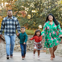 Hetal in pink and green dress with husband in plaid shirt, son in green shirt, and daughter in red top plaid skirt walking across a wooden bridge