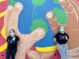 Abby Kietzman and Shannon Hill, RN, BSN wearing facemasks and standing outside, in front of a mural with three hands holding the planet Earth.