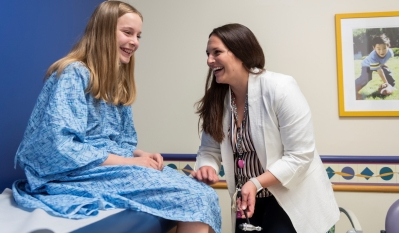 A teenage girl with blond hair sits on a clinic table while a clinician tests her knee reflexes.