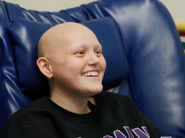 A teenage girl without hair smiles as she sits in a blue chair.