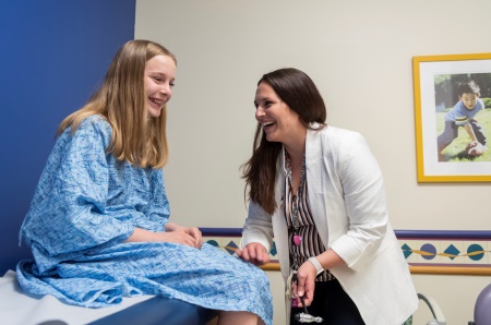 A teenage girl with blond hair sits on an exam table while a female clinician checks her knee reflexes.