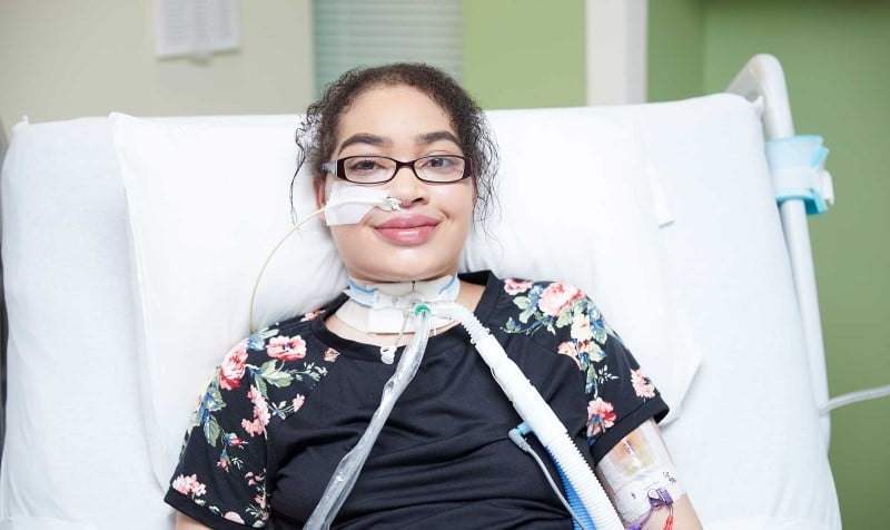 A teenage girl with a tube in her nose and in her neck smiles in a hospital bed.