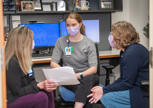 Three masked women sit together and talk in an office setting.