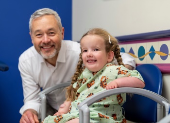 A young girl with red braided hair smiles while wearing a hospital gown. A male physician sits with her.