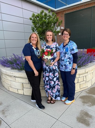 A three women pose together outdoors, the middle woman holding a vase of flowers