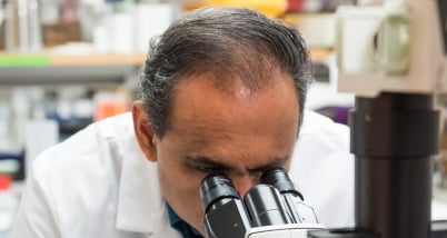 A man with brown skin and black hair looks into a microscope.