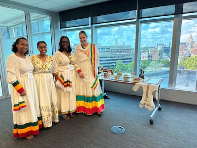 4 women in Traditional Ethiopian attire that is cream white with green, yellow, and red stripes on the ends of the skirt. 