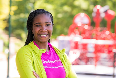 Children's Mercy Staff Nurse, Crystal, standing outside of the playground at Adele Hall. She is smiling and wearing a pink Children's Mercy t-shirt.