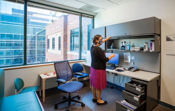 A masked, female medical professional opens a cabinet in an office.