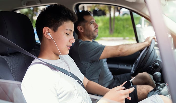 Teen boy sits in the passenger seat of the car with his dad listening to a phone with headphones.