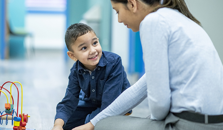 Young boy wearing a dark blue shirt sits on the floor next to a woman in a light blue shirt. They are playing with a toy on the ground.