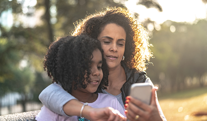 Mom hugs her preteen daughter and looks at a cell phone.