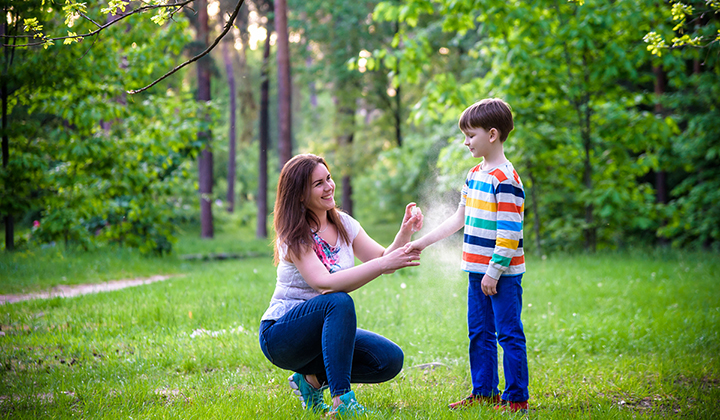 Woman sprays bug repellant on young child while outside in a woody area.