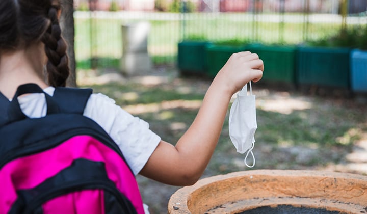 Young girl throws away a face mask in a playground trash can.