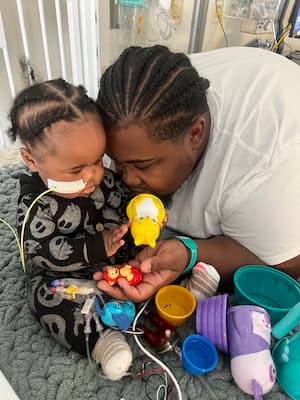 A dad and baby play with toys together. There are multiple tubes and bags attached to the baby’s body and hospital room equipment in the background. 