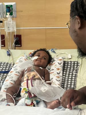 A baby in a hospital bed looks up at his dad whose profile is in the foreground. The baby has many tubes and wires attached to his body and there is more hospital equipment in the background. 