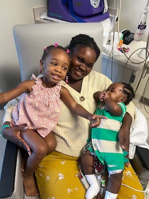 A mom smiles from a hospital chair as she holds a baby and a toddler. The baby has medical equipment and tubes peeking out of his onesie. The toddler is smiling and pointing to something out of frame. There is more medical equipment in the background.