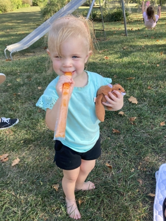 Geneva, a toddler with light blonde hair, enjoys a popsicle by her backyard swing set.