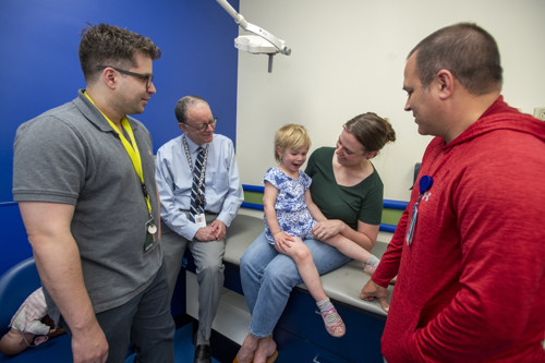  Geneva, a toddler in a blue and white romper, sits on her mom's lap as her craniofacial team looks on. They are in a clinic room at Children's Mercy.