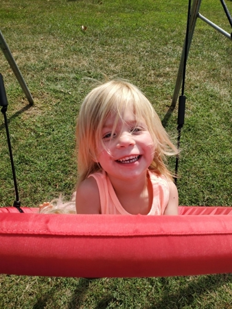  Geneva, a smiling, blonde-haired toddler, swings on a red disc swing.