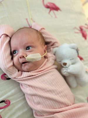 A baby in a pink onesie looks off camera. She has a small yellow tube taped to her face that goes into her nose. A white teddy bear is at her side, and her sheet has pink flamingos on it.