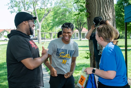 A group of students and adults interact in a park setting. The Black student at the middle of the photo is laughing.